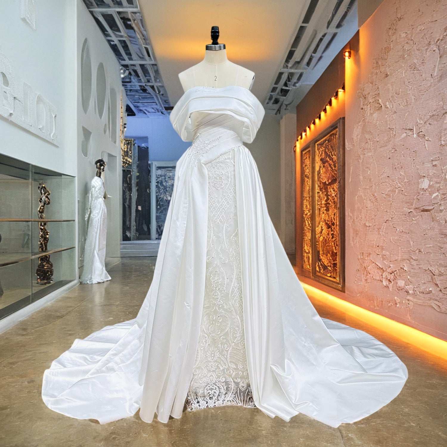 Ivory off-the-shoulder satin ball gown bridal dress with a split-front overskirt and a beaded lace column underskirt displayed on a mannequin in a modern showroom corridor.