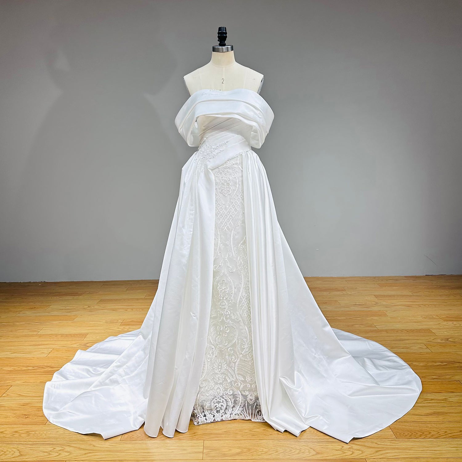 Front view of a premium white satin bridal gown showcasing the unique wide-cuffed off-the-shoulder portrait neckline and asymmetrical pearl-beaded waist detailing on a dress form.