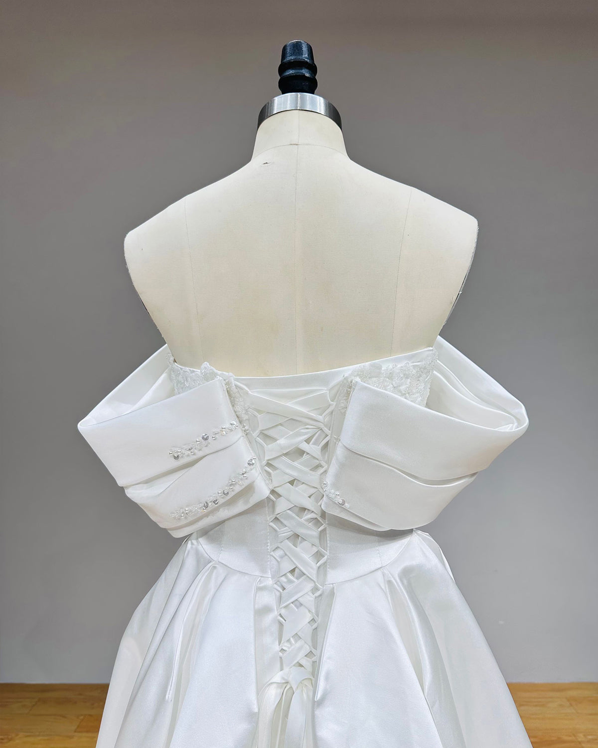 Close-up back view of a satin wedding dress showing the detailed lace-up corset closure, delicate pearl embroidery along the upper trim, and architectural cuffed sleeves.