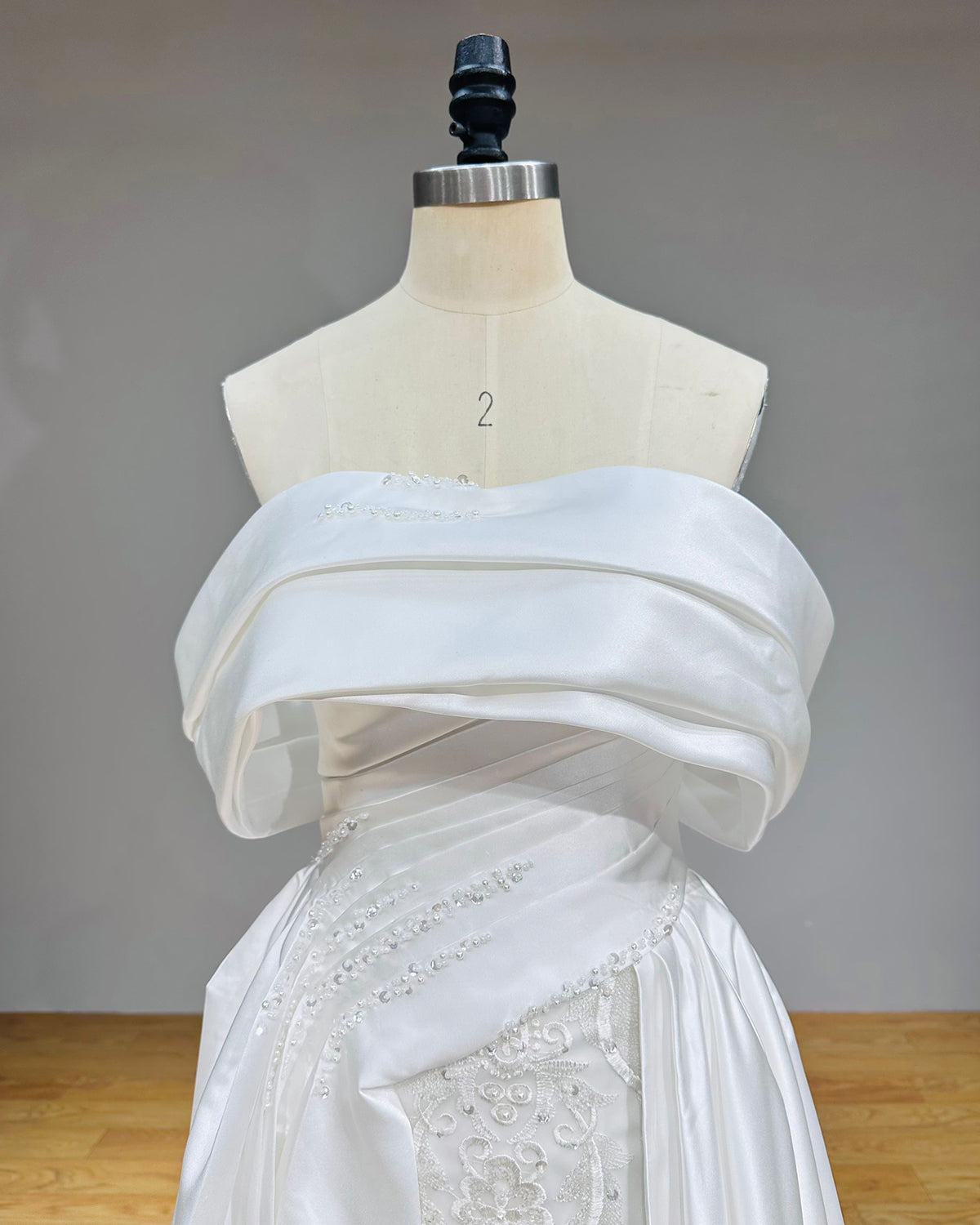 Detailed close-up of a structured off-the-shoulder wrap bodice on an ivory satin bridal gown highlighting diagonal pleating and trailing pearl embellishments.