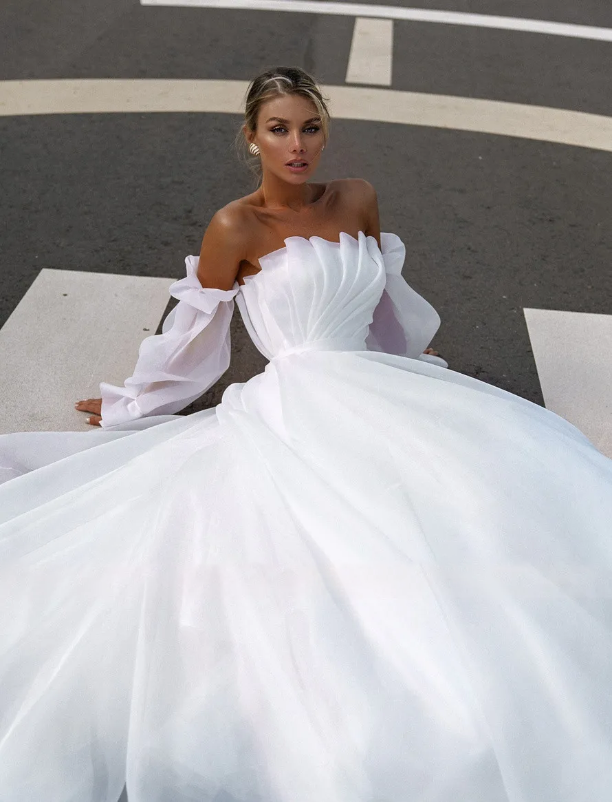 Bride sitting in a pleated A-line wedding dress with detachable puff sleeves, highlighting the modern boat neck silhouette.