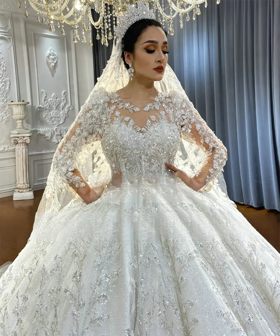Elegant bride posing in a shimmering long-sleeve ball gown bridal dress with a high illusion neckline, paired with a royal crystal tiara and a long lace-trimmed veil.