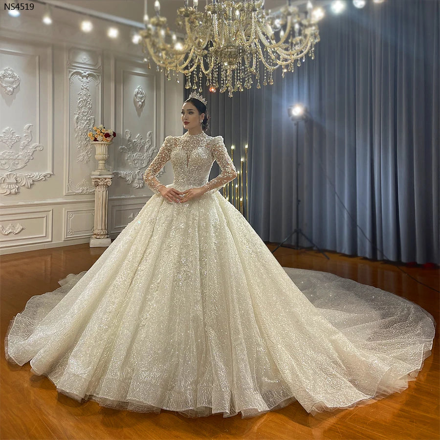 Full-length view of a bride wearing a new design luxury long sleeve ball gown wedding dress with a sheer high-neck bodice and an expansive sparkling skirt in a classic showroom.