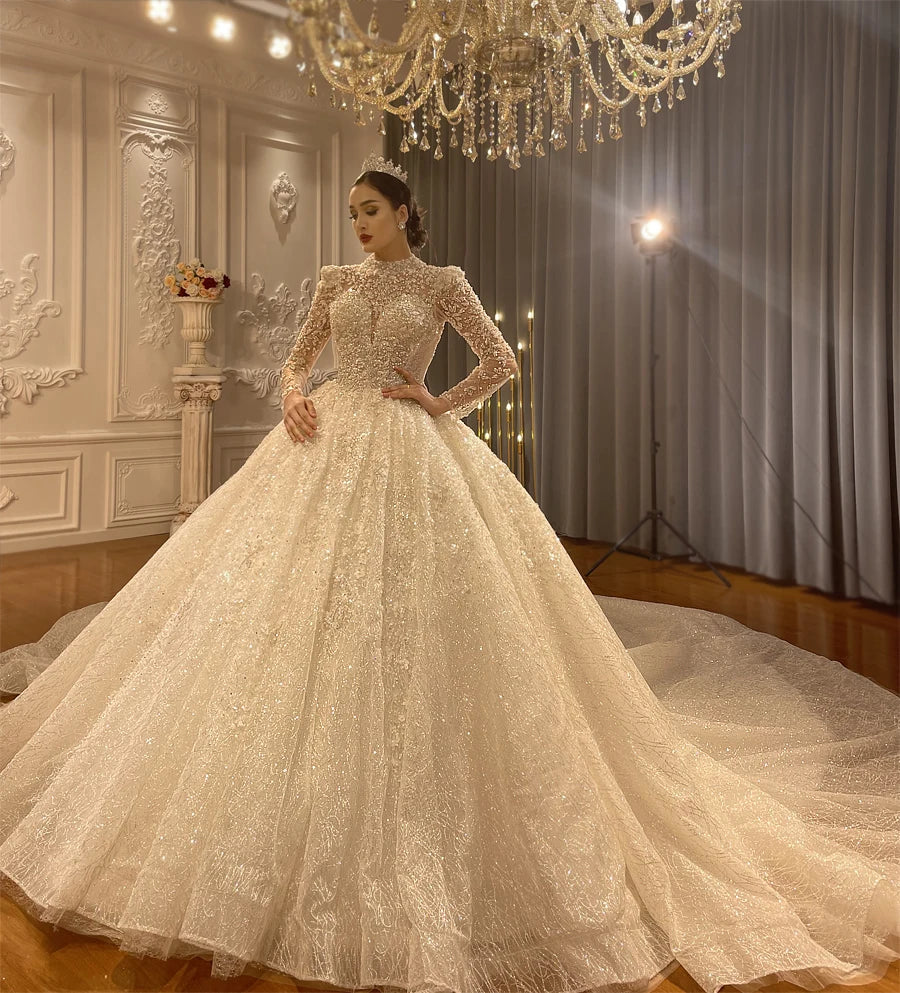 A regal bride showcasing a luxury sparkly ball gown wedding dress featuring long sleeves and a long cathedral train under a grand chandelier.