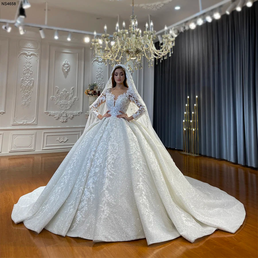 Full-length front view of a luxury long-sleeve ball gown wedding dress featuring an intricate floral lace bodice and a voluminous shimmering white skirt, showcased under a grand crystal chandelier in an elegant bridal showroom.