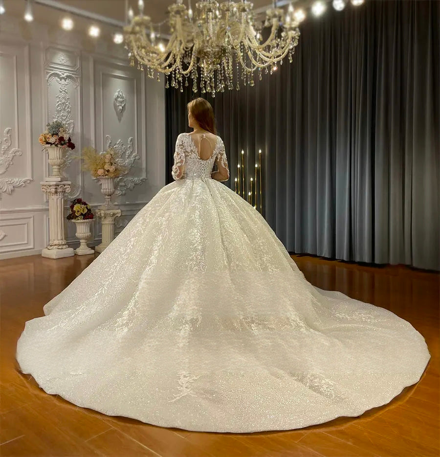 Rear perspective of an elegant bridal gown with long lace sleeves, highlighting a sophisticated keyhole back design and a majestic, expansive sparkling cathedral-length train spread across the floor.