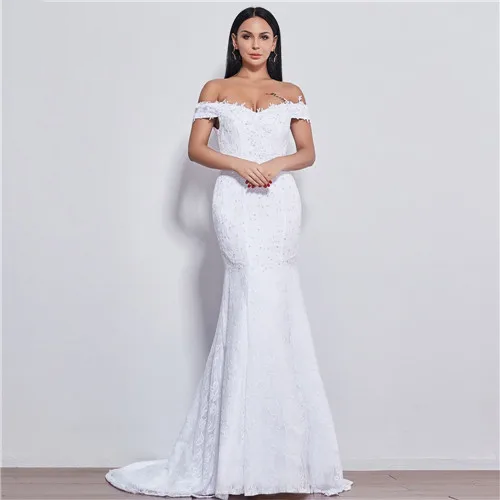 A beautiful bride posing in a white off-shoulder mermaid wedding dress with intricate lace detailing, holding a large floral bouquet.
