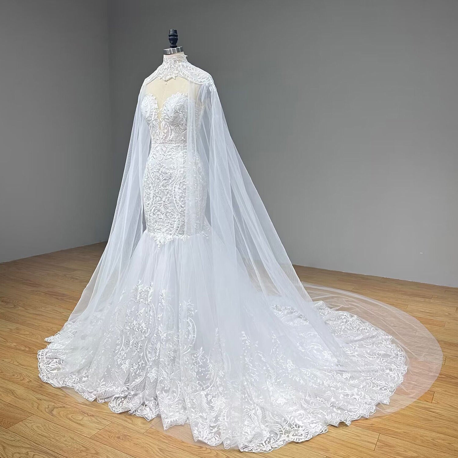 Three-quarter side profile of a form-fitting lace mermaid bridal gown on a dress form, displaying the intricate baroque applique patterns and a flowing sheer tulle cathedral cape.