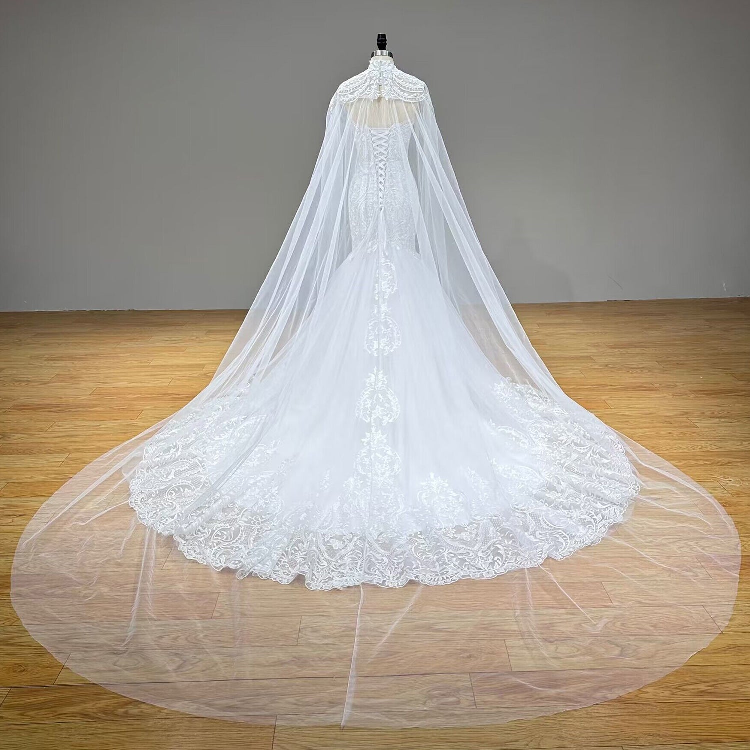 Full back view of an ivory tulle mermaid wedding dress on a mannequin, showcasing the lace-up corset closure and a long, dramatic sheer tulle bridal cape sweeping over a scalloped lace train.