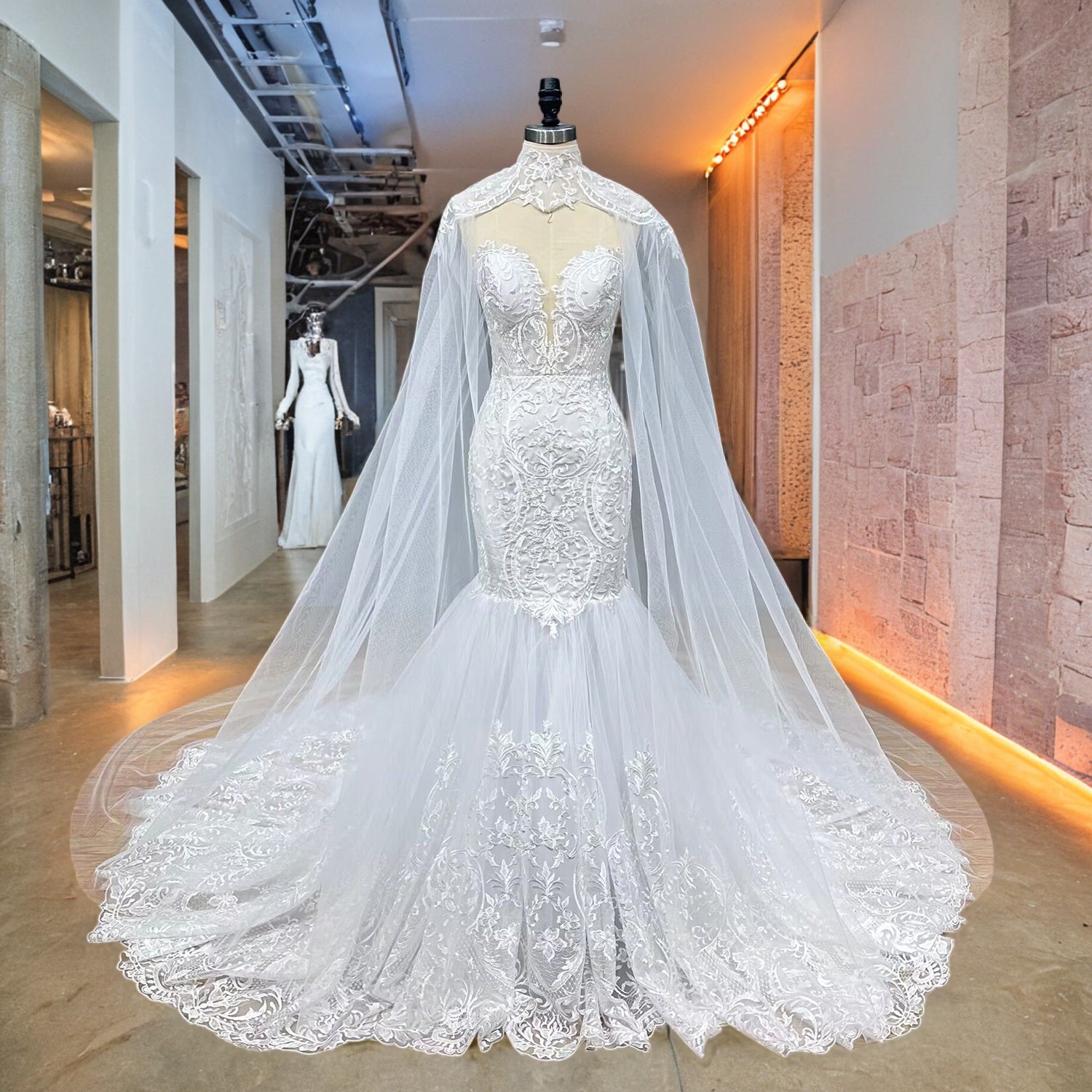 Full front display of an ivory tulle trumpet wedding dress with intricate lace appliques and a matching high-neck sheer capelet on a studio mannequin.