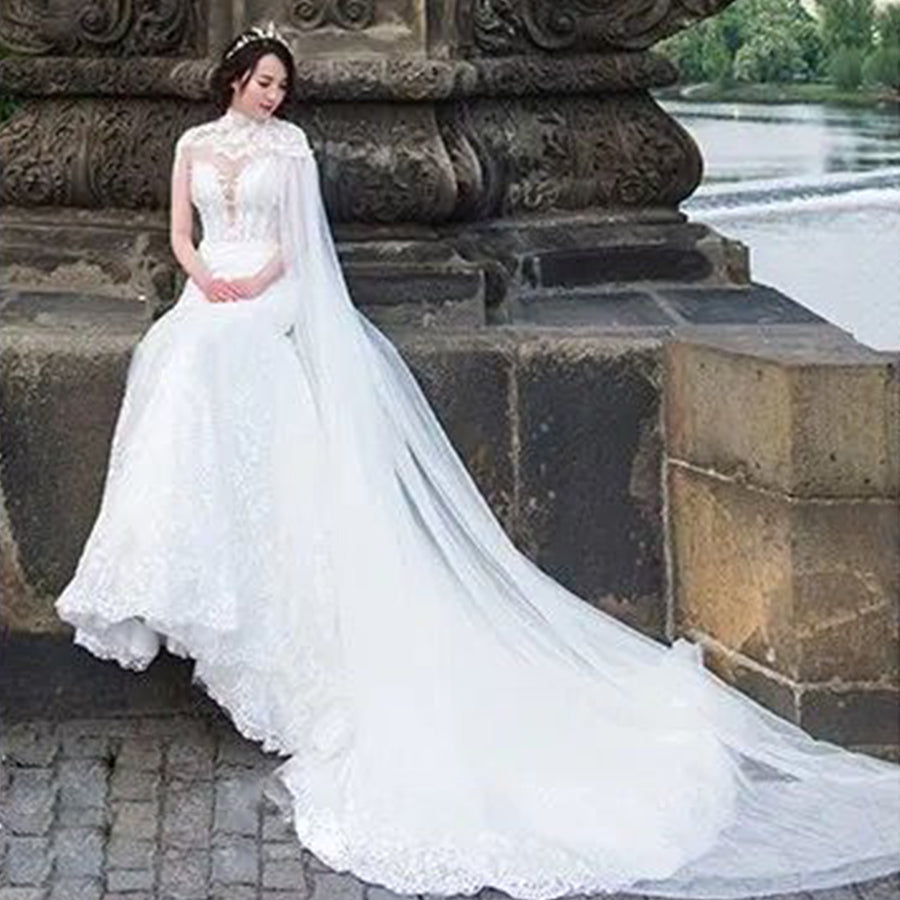 A bride sitting on a historic stone ledge by the water, showcasing the dramatic length of a sweeping sheer tulle bridal cape attached to a lace mermaid gown.