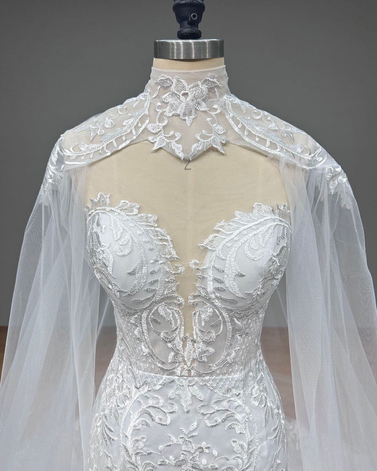 Detailed front view of a luxury bridal gown bodice on a dress form, displaying symmetrical baroque lace embroidery, sheer inserts, and a cutout capelet design.