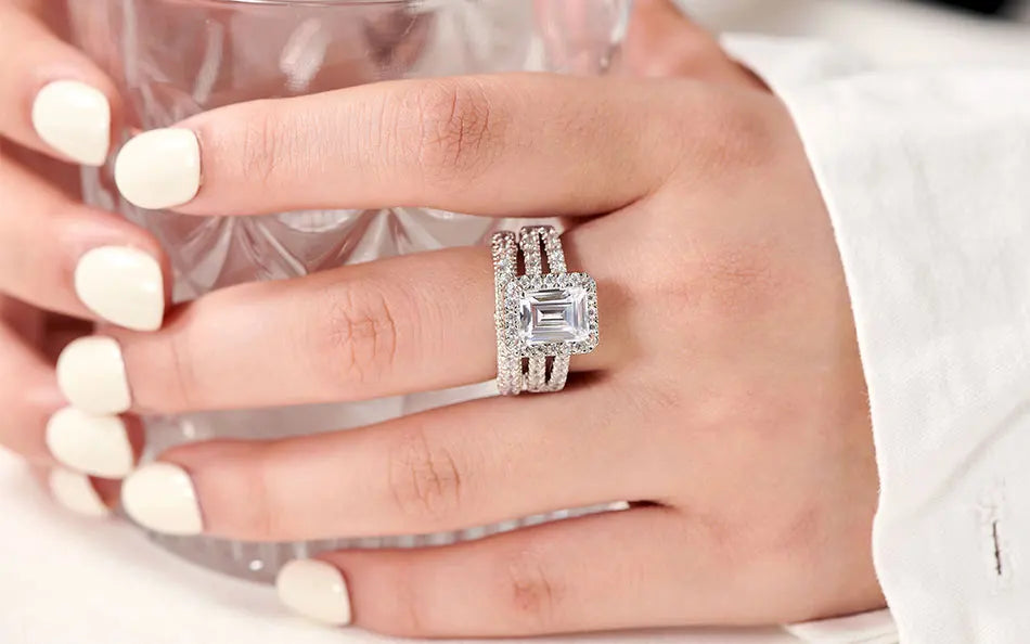Close-up of an emerald-cut cubic zircon engagement ring with a matching pave eternity band worn on a finger next to a textured glass.