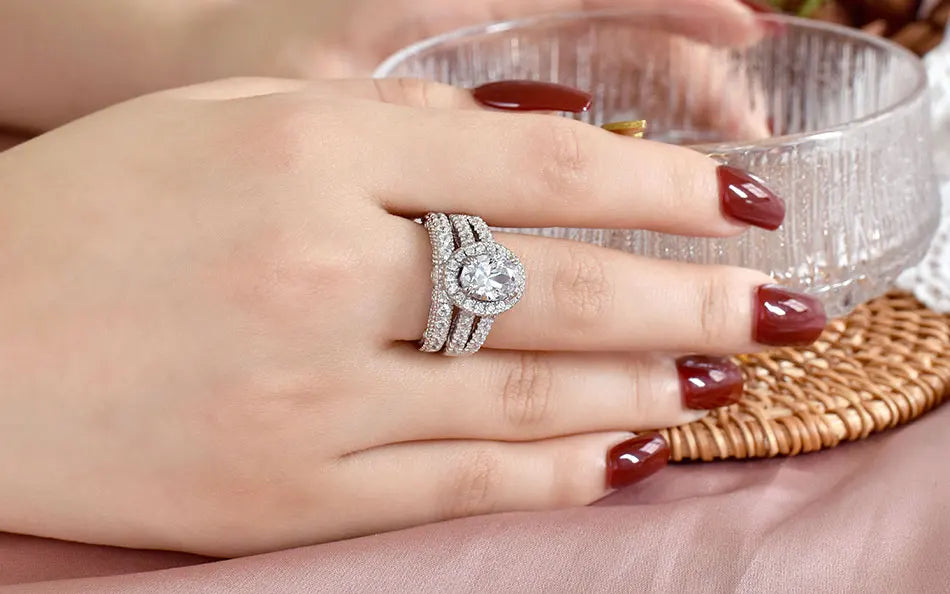 Oval-cut 5A cubic zircon halo engagement ring with a triple-band silver wedding set styled on a hand with dark red manicured nails.