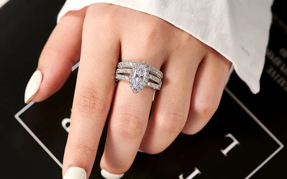 Oval-cut 5A cubic zircon halo engagement ring with a triple-band silver wedding set styled on a hand with dark red manicured nails.