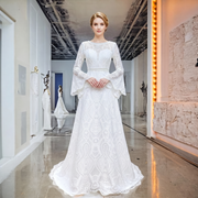Front view of an elegant model wearing an O-neck backless lace wedding dress featuring bohemian geometric patterns, long bell sleeves, and a see-through waist detail in a luxury showroom.