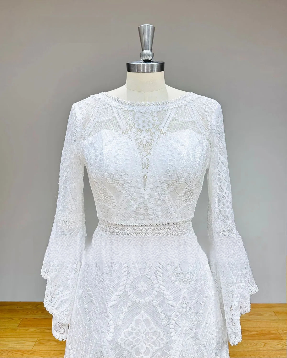 Close-up detail of a white lace bridal bodice on a mannequin, highlighting the high O-neckline, sheer see-through waist, and long bell sleeves with geometric lace.