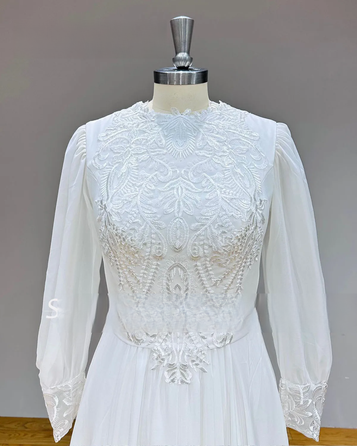 Close-up detail of the front bodice of a white chiffon wedding dress, featuring a high O-neckline with delicate floral lace embroidery and subtle beadwork.