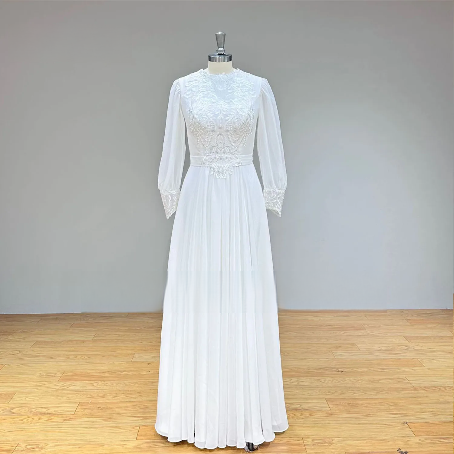 Full-length front view of a modest white long sleeve chiffon wedding dress on a mannequin, featuring a high O-neckline, intricate lace bodice appliques, and a flowing pleated skirt.
