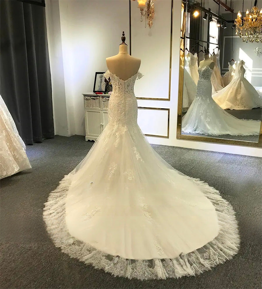 Angled back view of the elegant beach wedding dress on a mannequin, showcasing the sheer lace off-the-shoulder straps and the flared train.