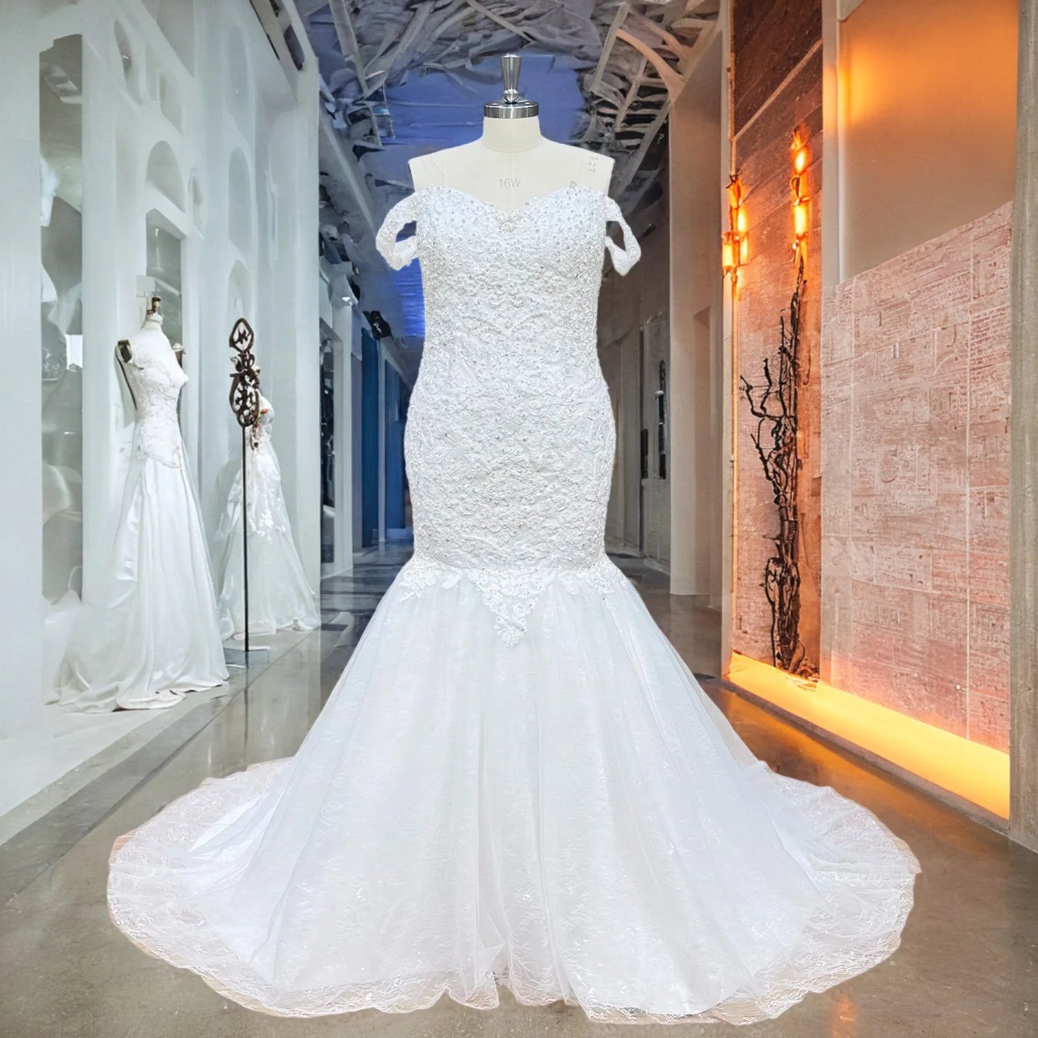 White off-the-shoulder mermaid bridal dress with dense floral lace embroidery and a dramatic tulle trumpet skirt displayed on a showroom mannequin.