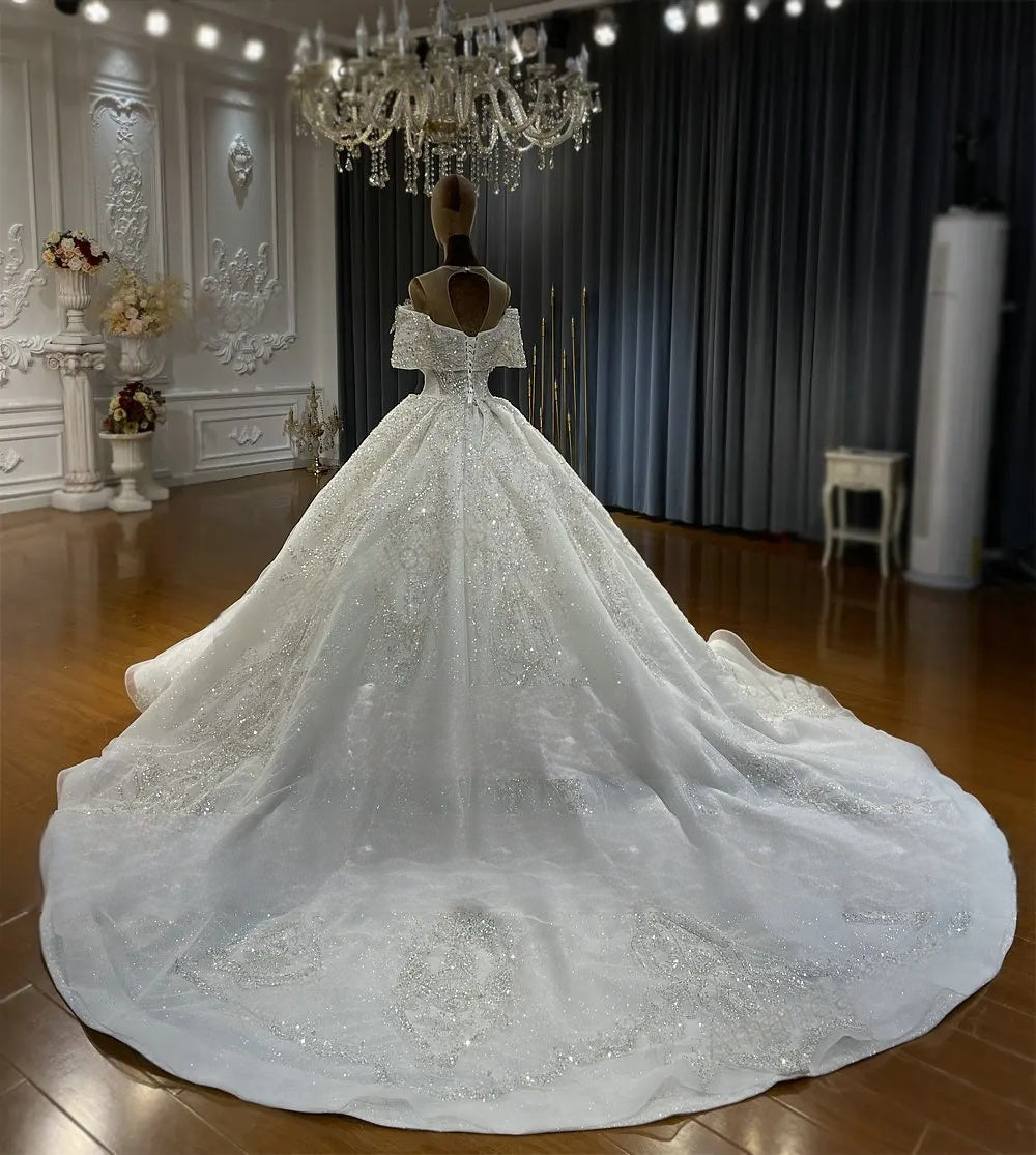 Rear view of a beautiful off-the-shoulder wedding dress on a mannequin, showcasing the elegant lace-up corset back and a massive shimmering crystal-beaded train.