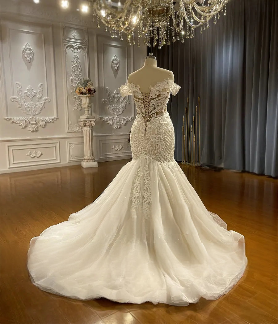 Back profile of the lace bridal gown, emphasizing the dramatic flare of the train and the intricate texture of the beaded lace overlay.
