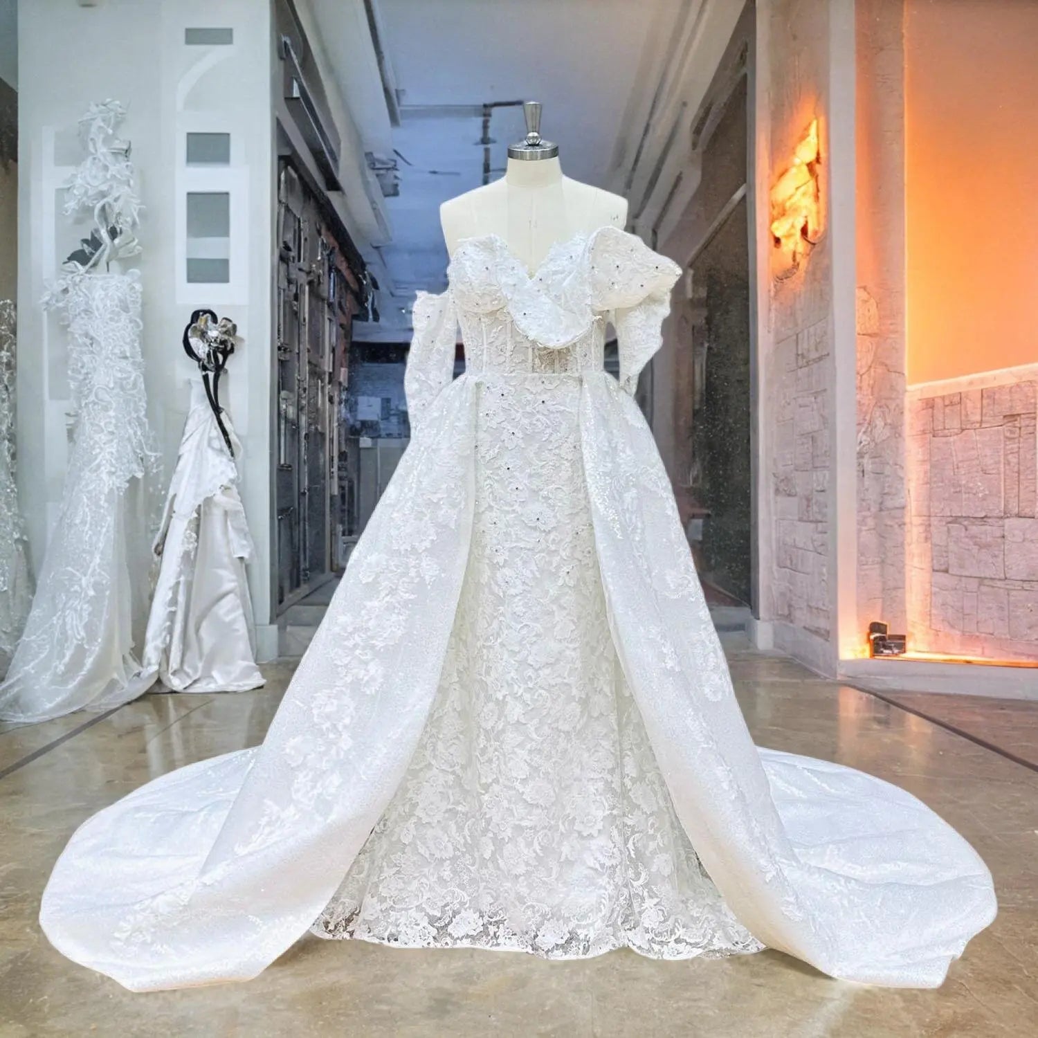 Front view of an off-shoulder 2-piece mermaid wedding dress on a mannequin, featuring an intricate beaded lace bodice and a dramatic, voluminous detachable overskirt train in a luxury bridal boutique.