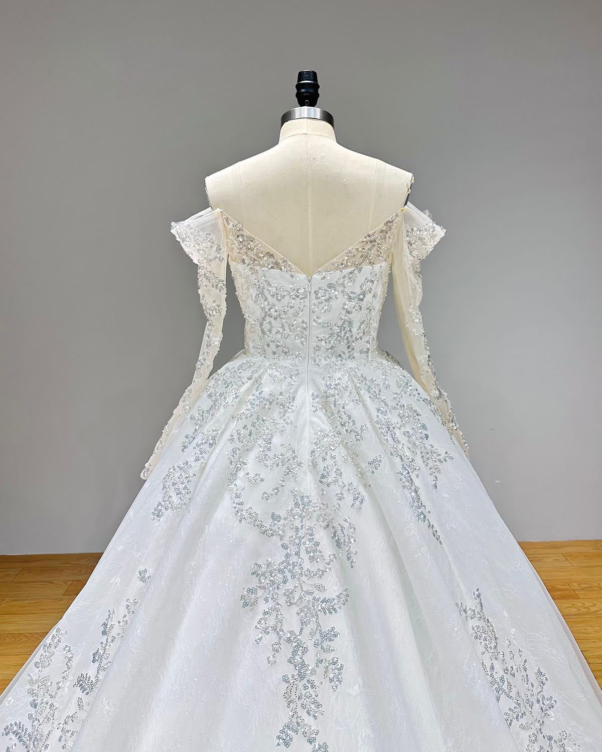 Close-up back bodice detail of an off shoulder ball gown wedding dress on a mannequin, highlighting the open V-back design, sheer long sleeves, and exquisite floral beading with pearls.