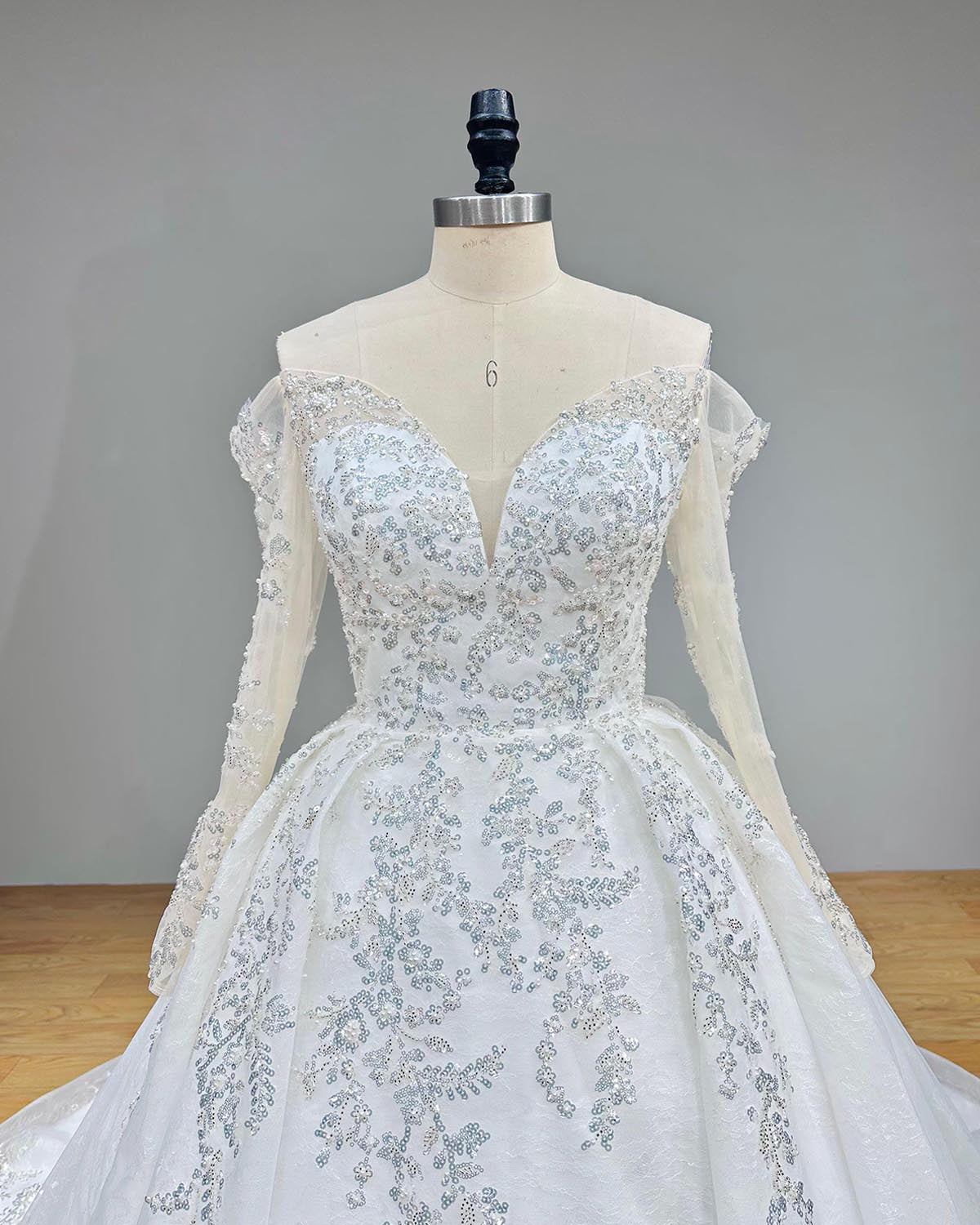 Close-up front bodice view of a luxury wedding gown on a mannequin, detailing the illusion sweetheart neckline, off shoulder sheer long sleeves, and heavy silver beading and pearls.