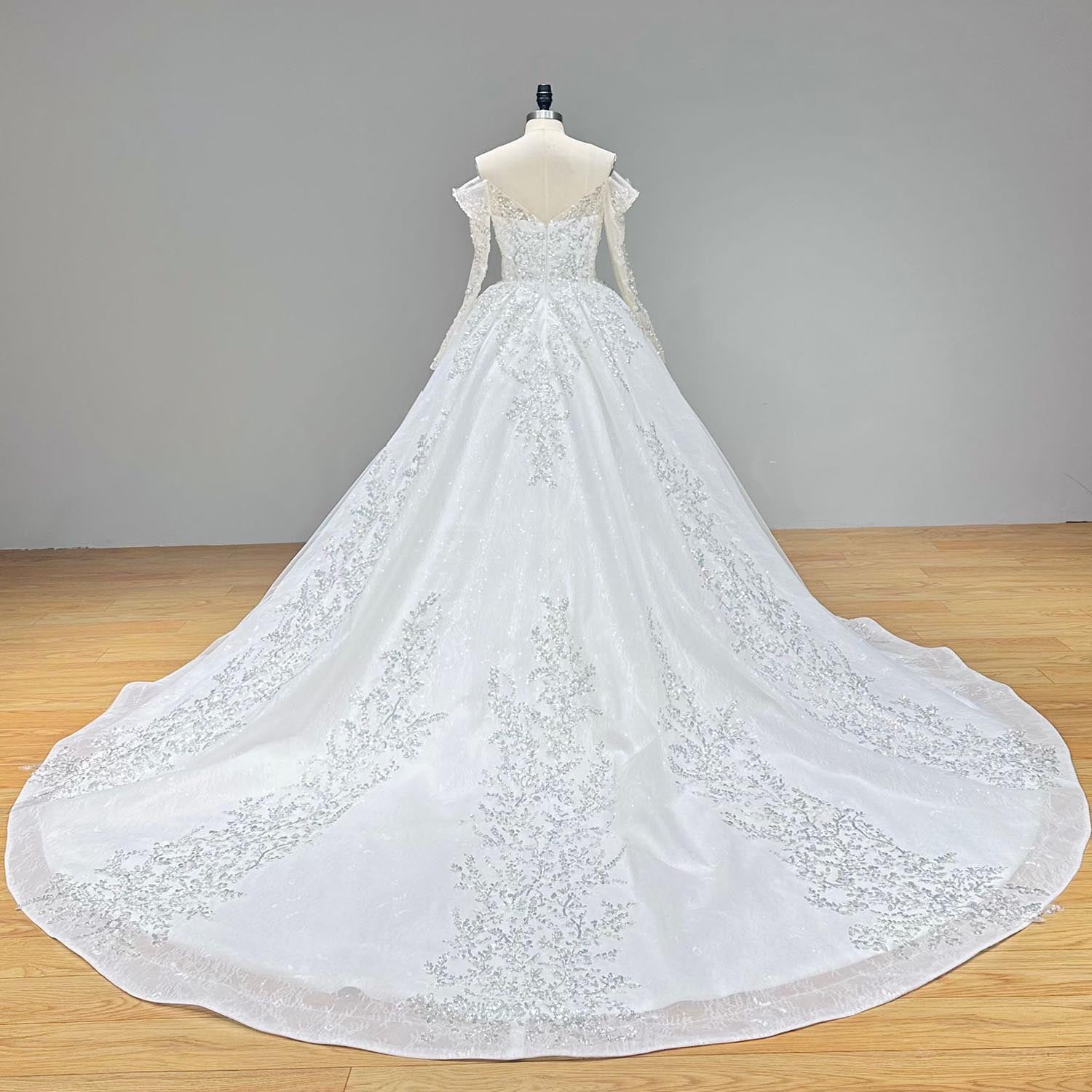 Dramatic full back view of an off shoulder ball gown wedding dress on a mannequin, displaying the delicate long sleeves and a sweeping, wide train heavily adorned with intricate beading and pearls.