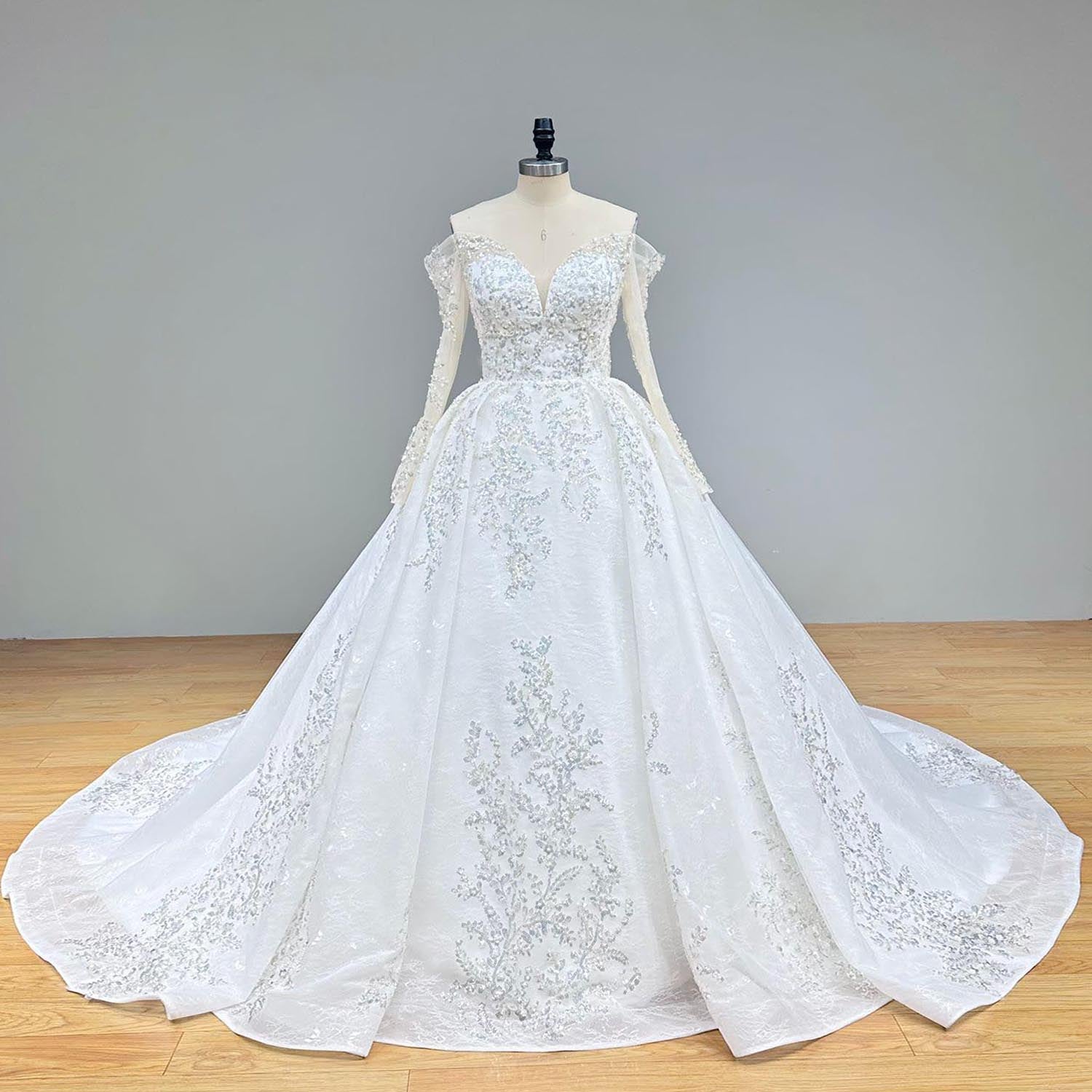 Classic full front view of an off shoulder ball gown wedding dress on a mannequin in a studio setting, showcasing the sheer long sleeves and cascading floral beading and pearls over a voluminous skirt.