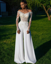 Full-length front view of a model standing outdoors wearing an off shoulder lace applique jumpsuit wedding dress with long sheer sleeves, a glittering sequined bodice, and fluid white wide-leg trousers.