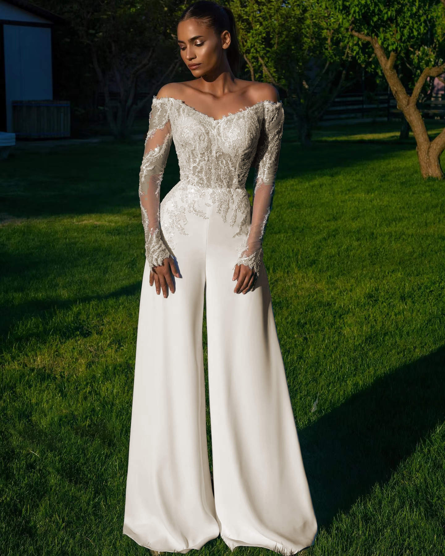 Full portrait shot of the white off shoulder bridal pantsuit, highlighting the intricate floral lace applique trailing down the sheer long sleeves and high-waisted trousers.