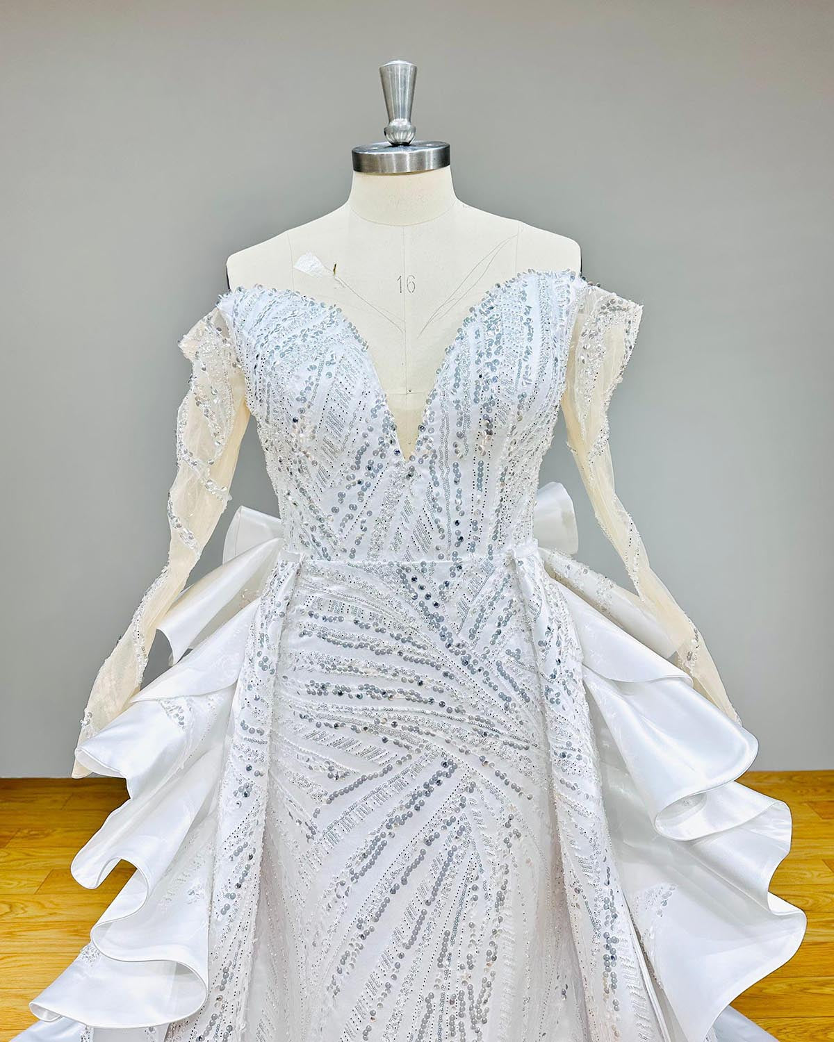 Close-up bodice detail of an off-shoulder mermaid wedding dress on a mannequin, focusing on the sheer illusion neckline, dense hand-beaded lace, and long sleeves.