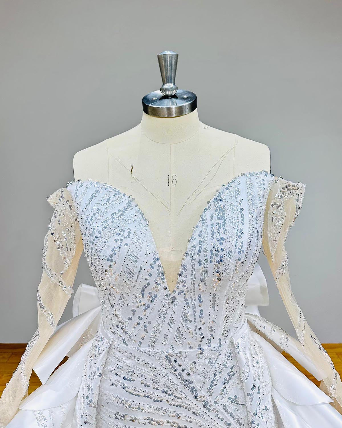 Close-up bodice detail of an off-shoulder mermaid wedding dress on a mannequin, focusing on the sheer illusion neckline, dense hand-beaded lace, and long sleeves.