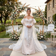 A smiling bride wearing an off-the-shoulder long sleeve mermaid wedding dress with beaded lace appliques standing under a wooden driftwood arch at a beach wedding ceremony.