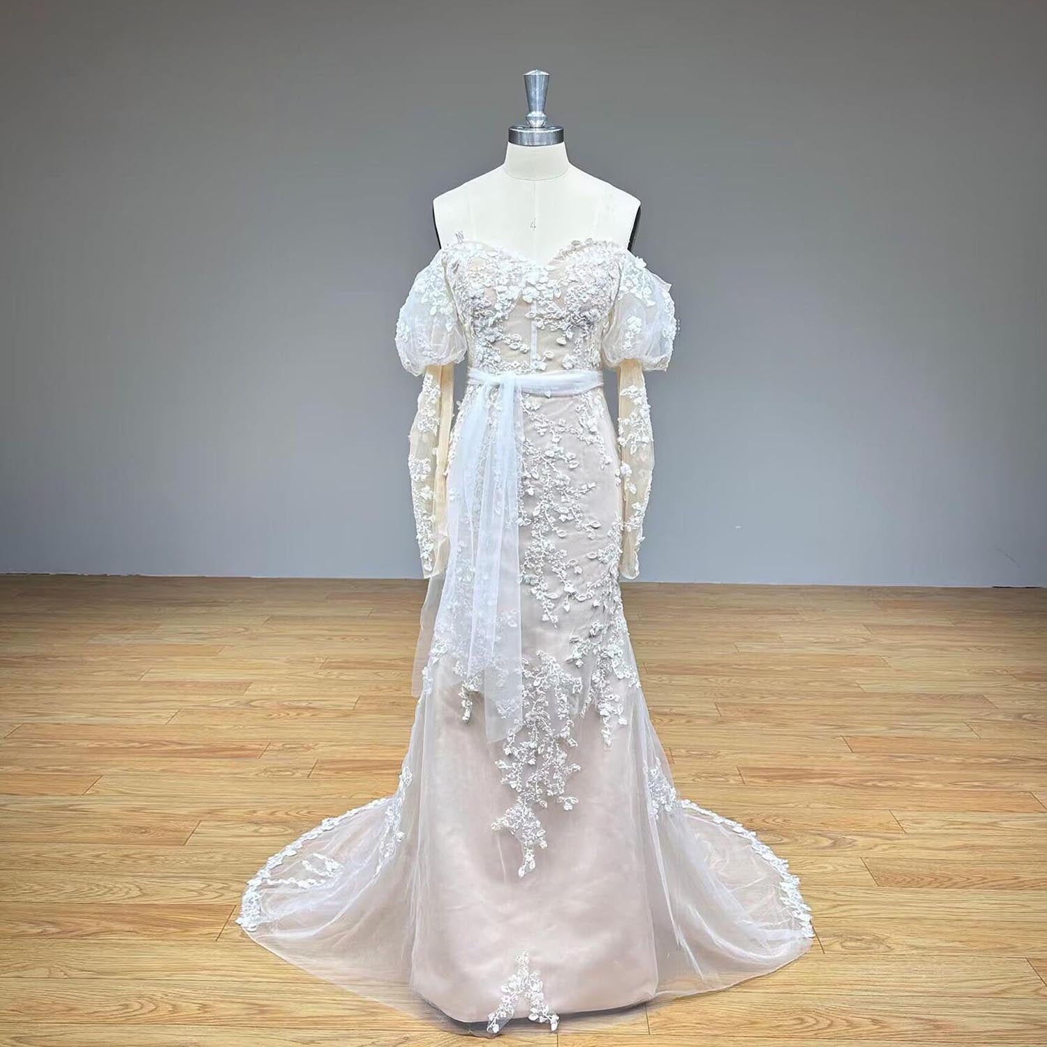 Studio product photography of a luxury vintage-style off-the-shoulder long sleeve lace trumpet bridal gown on a mannequin against a neutral gray wall backdrop.