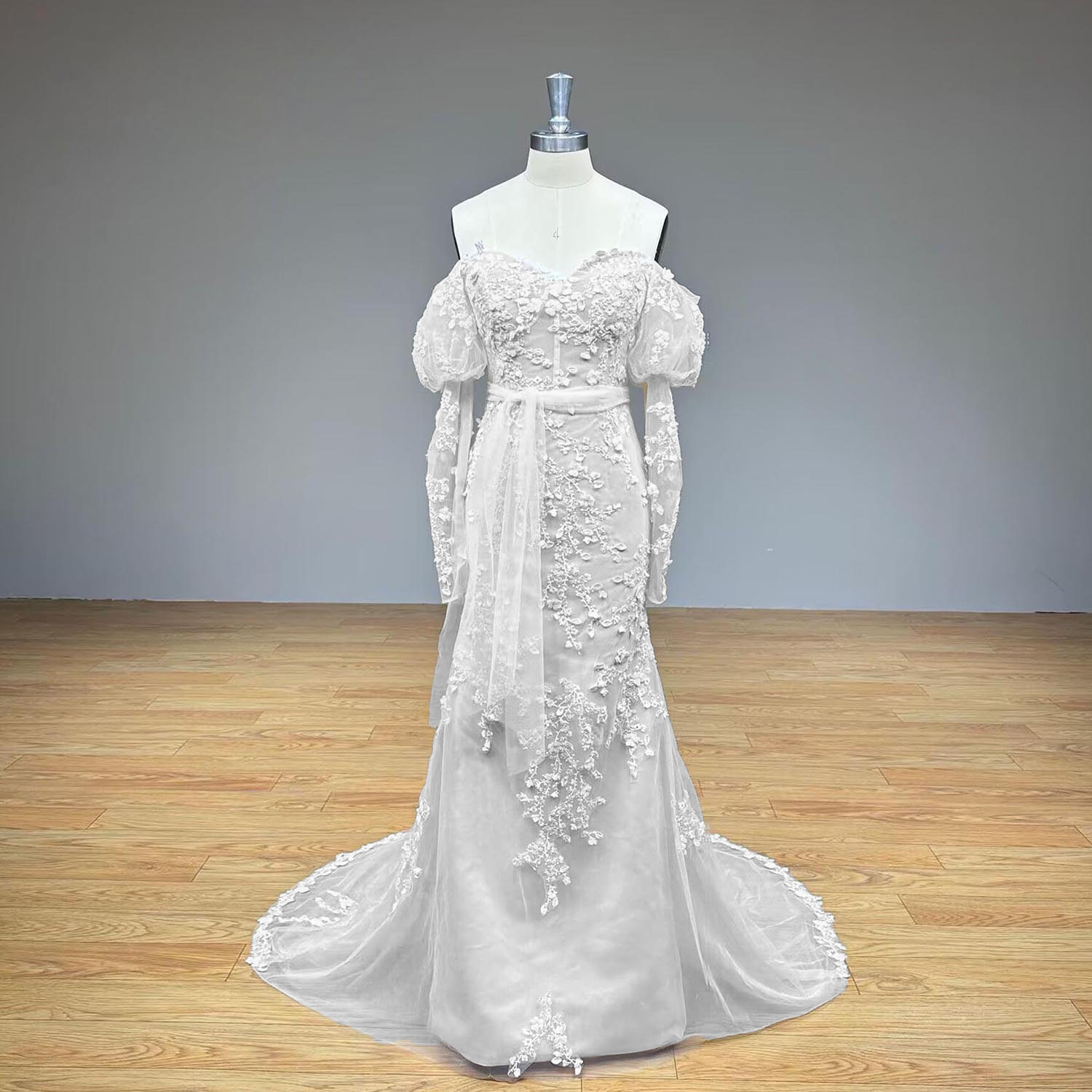 Studio product photography of a luxury vintage-style off-the-shoulder long sleeve lace trumpet bridal gown on a mannequin against a neutral gray wall backdrop.