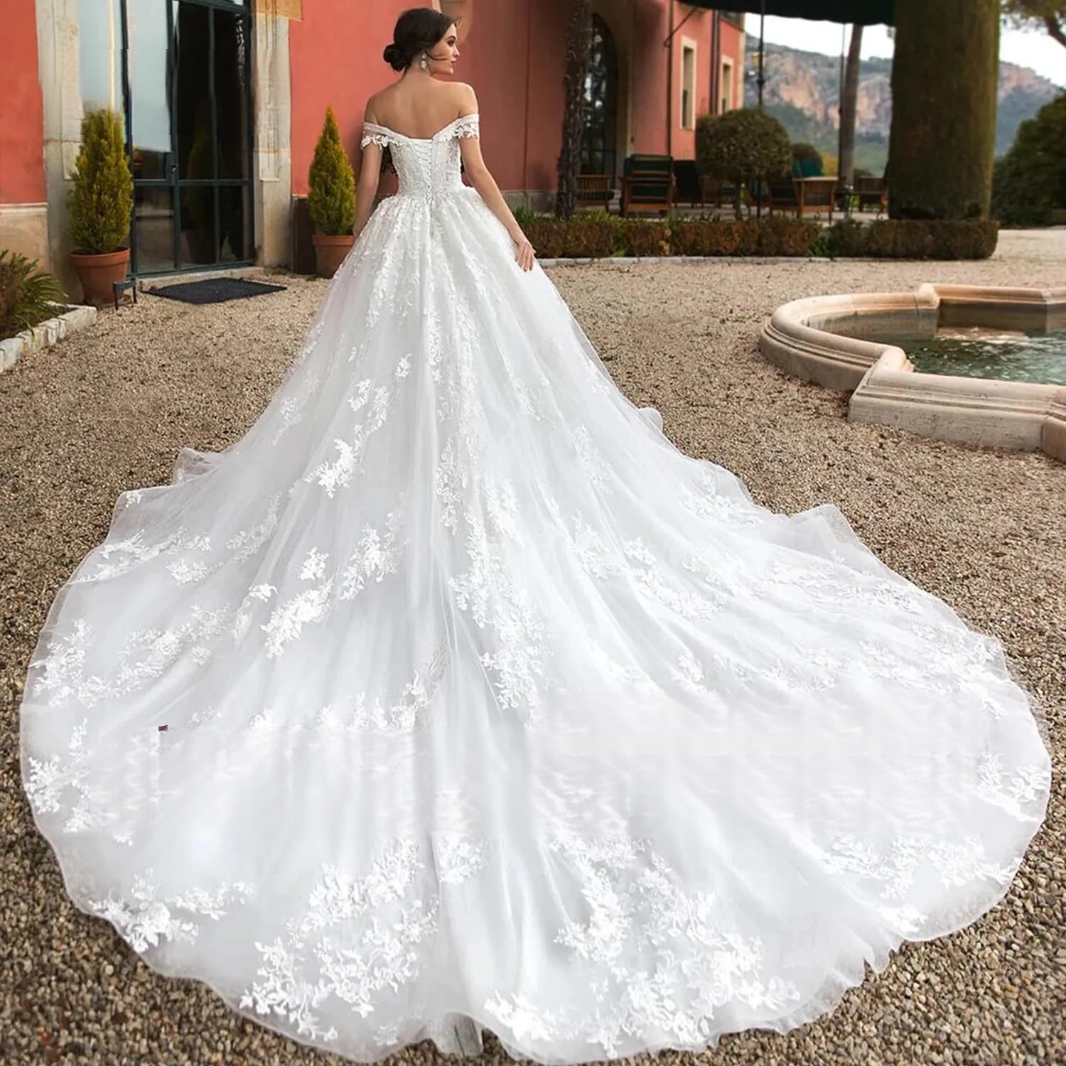 Outdoor back view of a bride wearing the off-shoulder sequin beaded ball gown wedding dress, showing the elegant lace-up back and the dramatic sweep of the long lace train.
