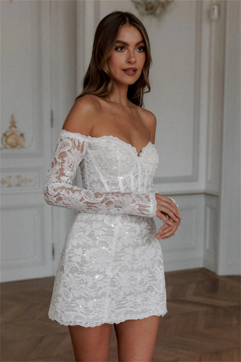 Side-facing profile of a short white lace bridal gown showcasing form-fitting off-shoulder long sleeves and a cleanly tailored mini sheath skirt line.