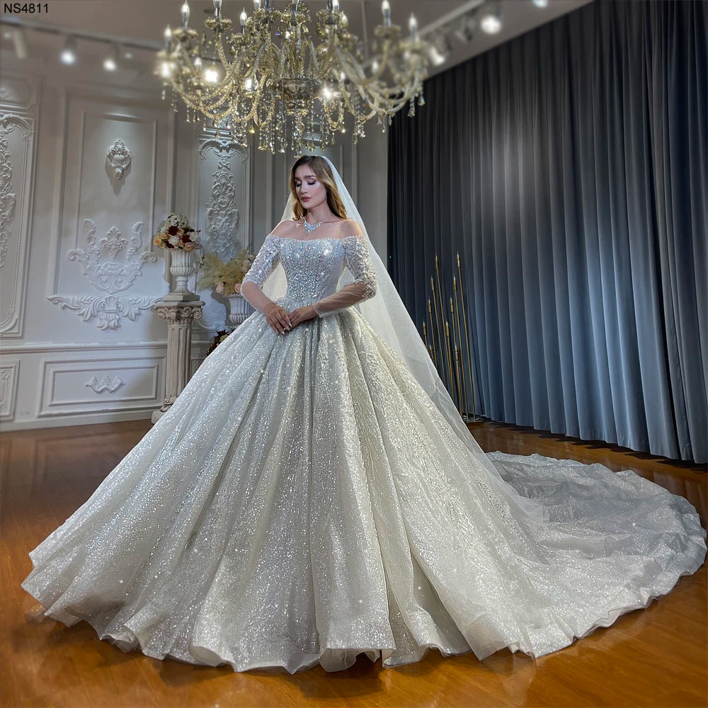 Full-length side perspective of an elegant off-the-shoulder long sleeve ball gown wedding dress, featuring a massive sparkling sequin train spread across a luxury bridal showroom.