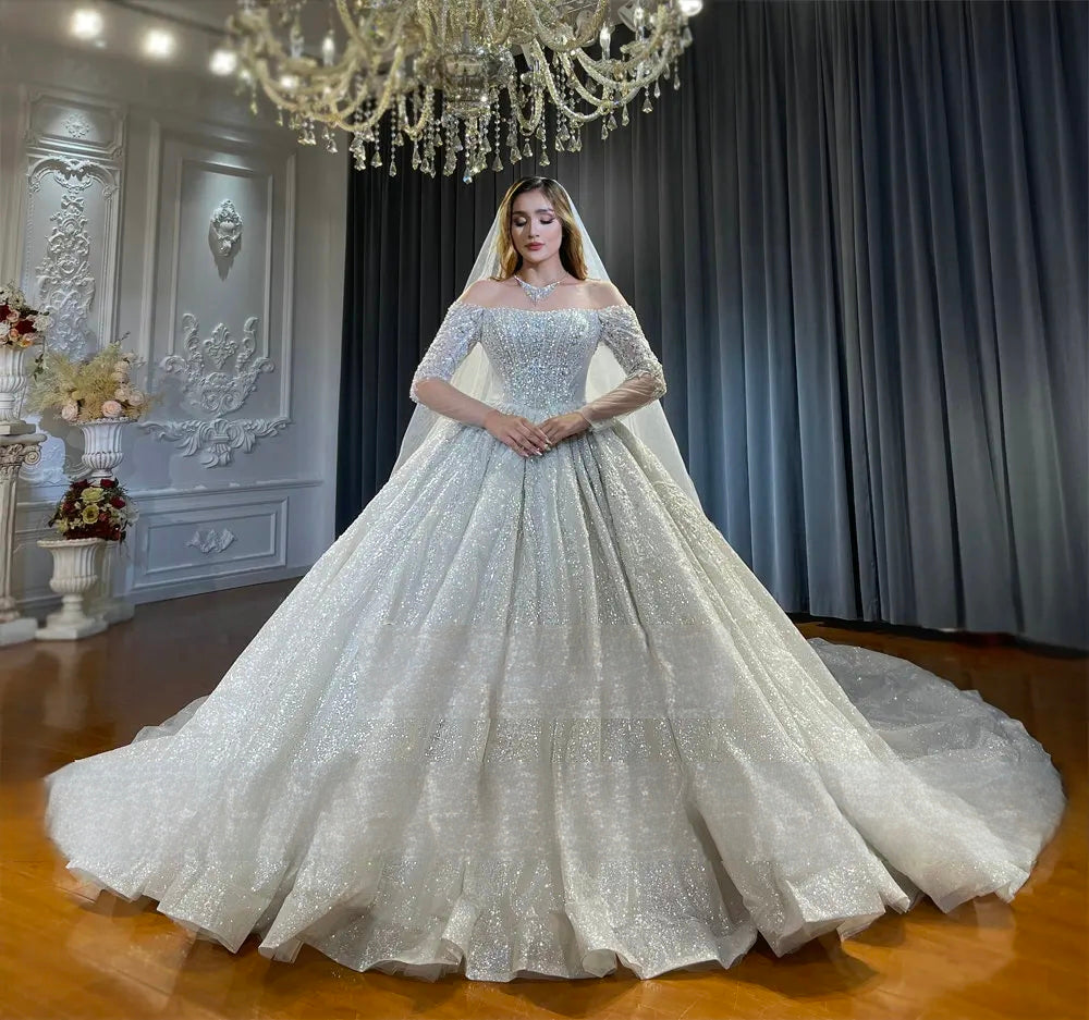 Front view of a stunning off-the-shoulder long sleeve bridal gown with a voluminous, fully-sequined sparkly ball gown skirt and a flowing sheer veil.