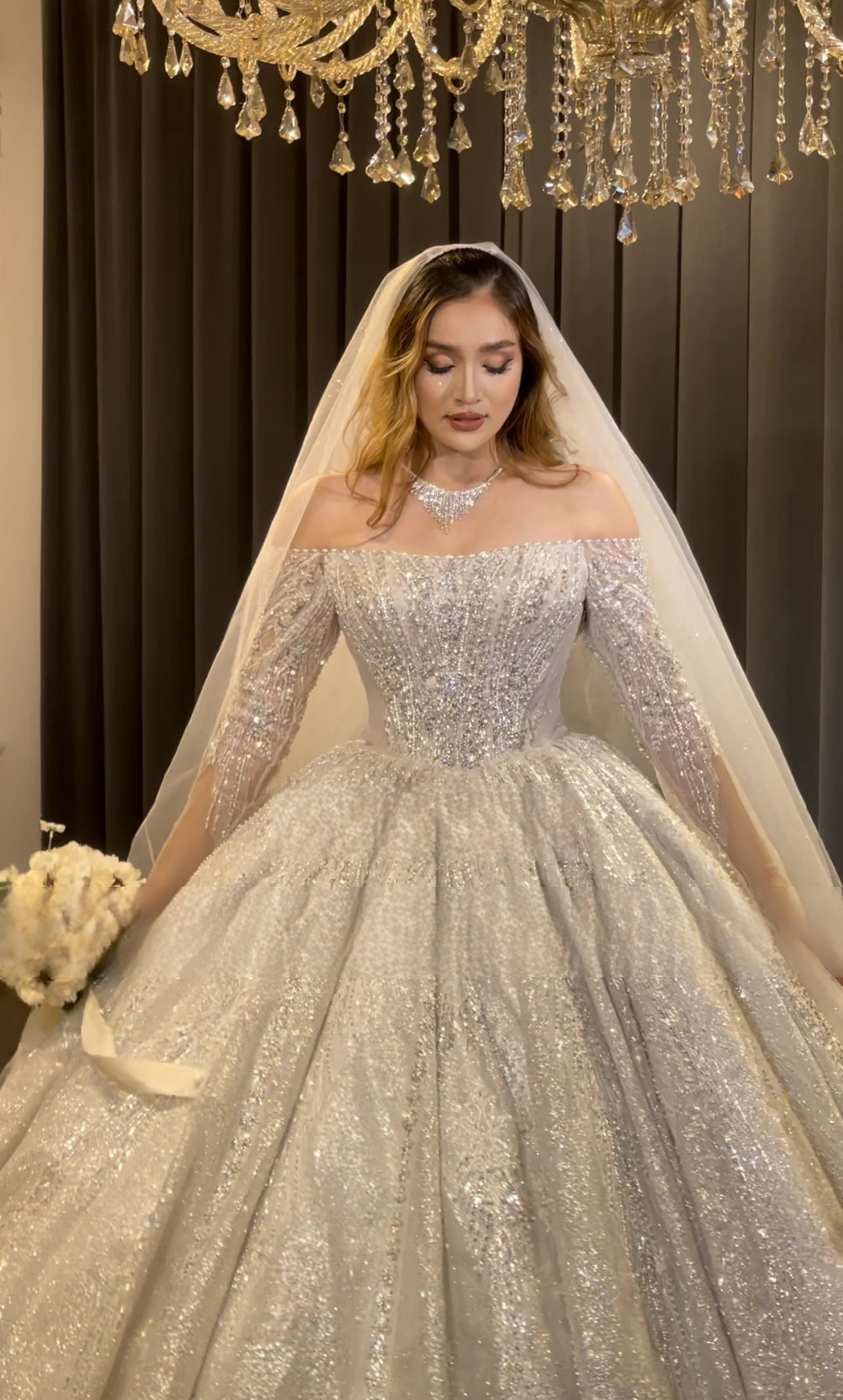 Beautiful bride posing in a glamorous off-the-shoulder long sleeve ball gown wedding dress completely covered in radiant crystal and sequin embellishments.