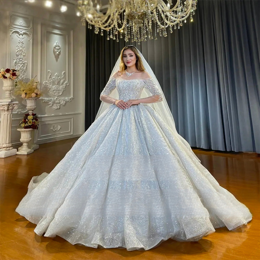 Front view of a stunning off-the-shoulder long sleeve bridal gown with a voluminous, fully-sequined sparkly ball gown skirt and a flowing sheer veil.