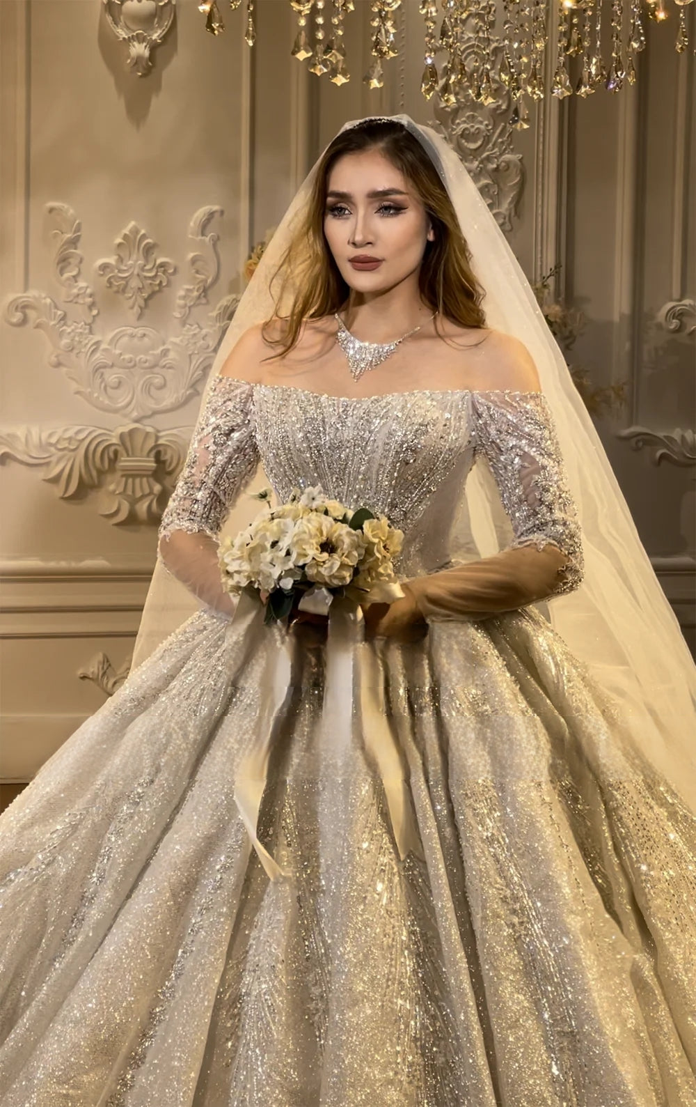Detailed front close-up of an elegant off-the-shoulder bridal gown showcasing the heavily beaded neckline, sparkling illusion sleeves, and bridal jewelry.
