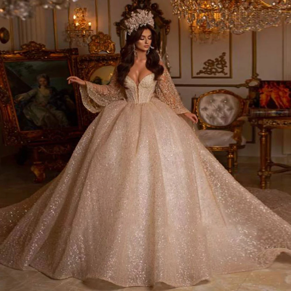 Dramatic off-the-shoulder ball gown wedding dress with a plunging V-neckline, heavily crystal-beaded corset bodice, and a voluminous shimmering glitter tulle skirt.