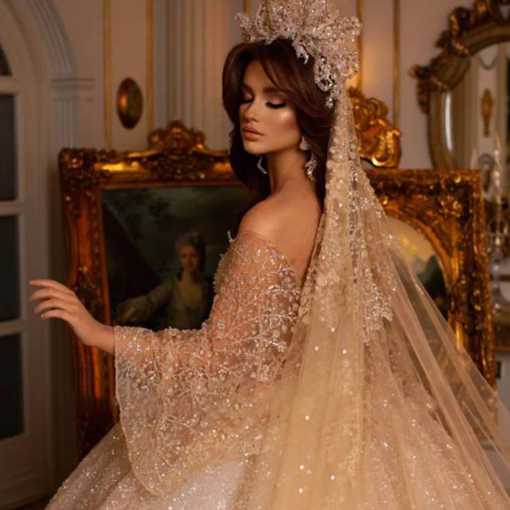 Romantic soft blush pink bridal ball gown featuring white crystal-beaded off-the-shoulder sleeve bands and a layered pastel pink shimmering tulle skirt.