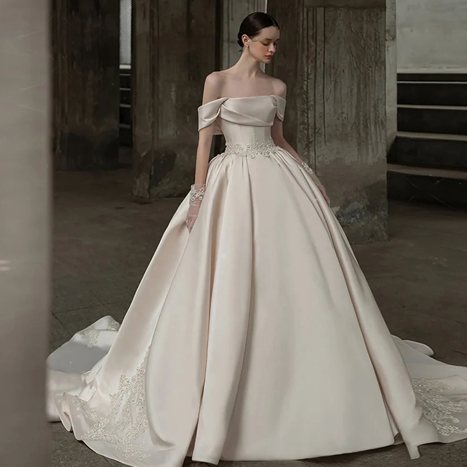 High-fashion bride wearing an ivory off-the-shoulder satin ball gown wedding dress with a pearl-embroidered basque waistline in an industrial studio setting.