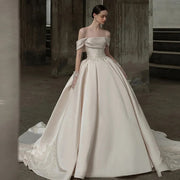 High-fashion bride wearing an ivory off-the-shoulder satin ball gown wedding dress with a pearl-embroidered basque waistline in an industrial studio setting.
